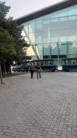 Two people, one wearing a dark shirt and grey pants, the other a brown shirt and patterned pants, walk away from the camera on a paved walkway. They are walking towards a large building with a glass facade. To their left are trees and a blue train-like vehicle. The sky appears to be reflecting in the building's glass.