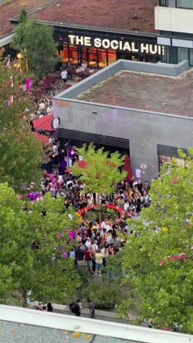 An aerial view shows a large outdoor gathering of people. The crowd is concentrated around a central area with trees and some red decorative elements, possibly an archway. Many individuals are standing and interacting. Some are wearing casual clothing. In the background, a building with the sign "THE SOCIAL HUB" is visible. There are also trees with green foliage, and some pink and purple lighting effects are present within the crowd. The scene appears to be an event or party taking place during daylight.