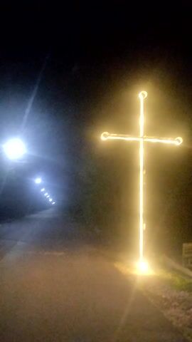 This video shows a dark outdoor environment, possibly a pathway or tunnel, illuminated by artificial lights. In the foreground, a bright white light source is visible on the left. In the middle ground and background, a series of smaller white lights are spaced out along a path, receding into the distance. Above these lights, a large, illuminated cross-shaped structure is visible, emitting a warm yellow glow. The cross appears to be made of lights or illuminated material and is positioned horizontally across the frame. The overall impression is of a decorated or marked path at night.