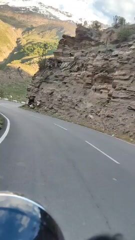 The video shows a view from a motorcycle as it travels along a winding mountain road. The road is paved and has white lane markings. To the right of the road is a steep, rocky cliff face. To the left, the terrain slopes upwards, covered in green grass and dotted with trees. In the distance, snow-capped mountains rise against a partly cloudy sky. Several orange and yellow safety barriers are visible along the left side of the road. The rider's helmet is partially visible in the lower foreground, with a reflection of the sky and mountains on its surface. The sun appears to be casting shadows and highlighting the landscape.