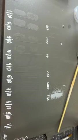 The video displays a dark surface, possibly a workbench, with various markings and substances. On the left side of the surface, there are vertical rows of faded markings that appear to be handwritten in white, with some numbers visible such as "1/10", "2/10", "3/10", "4/10", "5/10", "6/10", "7/10", "8/10", "9/10", and "10/10". Adjacent to these markings, there are faint streaks of what looks like paint or a similar substance.

Further to the right, there are columns of handwritten text in white, with some characters resembling Russian or Cyrillic letters. These appear to be labels for different experiments or mixtures. For example, one column has labels like "Лез", "шар", "гл", "Лез", "шар", and "гл". There are also some scattered dots and a visible "X" and "Г" mark on the surface.

In the lower right portion of the frame, a brush with a light-colored handle and bristles is present. Scattered on the surface near this brush is a powdery substance, possibly pigment or powder, which is light blue or grey in color.

In the upper right portion of the video, a small digital scale is visible, along with a clear plastic container holding some small items. Further to the right, there are several white cylindrical containers with colored lids, and some clear plastic cups with handwritten labels.

The overall impression is that of a workspace where experiments or tests involving pigments, mixtures, and application techniques are being conducted. The lighting suggests an indoor environment. There are no people visible in the frame.