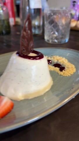 The video shows two plates on a dark brown table with a plate holding a white flan decorated with strawberry slices and dark red jam on top with a chocolate decoration. There is another plate in the background, similar to the first. In the foreground, there is another flan of a darker, caramel-like color, with similar toppings. A silver spoon lies next to the flan. A bottle of water, glasses, and decorations are in the background. The camera is focused on the plate with the caramel-like flan.