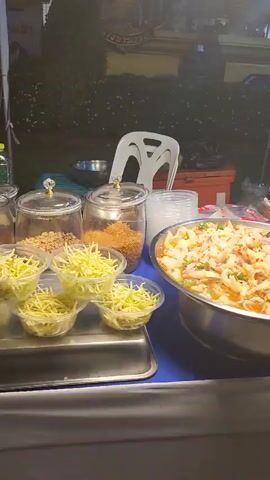 The video pans across a food stall at night. In the foreground, a large metal bowl is filled with a dish that appears to be spicy seafood salad, possibly containing squid and shrimp, mixed with herbs and chili peppers. To the left of this bowl are smaller plastic containers holding shredded green vegetables, likely mango and possibly lime or green papaya. Another jar behind these contains a reddish-brown granular substance, possibly fried shallots or chili flakes. Further to the right, another large metal bowl contains a similar spicy seafood salad, this one appearing to have more shrimp. A serving spoon is visible in both bowls. Behind the food, a white plastic chair is visible. A person, wearing a white top and a purple apron, stands at a table to the right, seemingly preparing food. Various containers and a large water dispenser are also on the table. The background is dark and out of focus, suggesting an outdoor setting like a night market. The lighting is dim, typical of nighttime street food vendors.