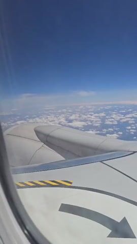 The video shows a view from an airplane window. The camera pans across the wing of the airplane, which is white with some markings. Below the wing, there is a layer of white clouds, and above the clouds is a clear blue sky. The sun is shining, and there are some glints of light on the wing. In the distance, the curvature of the earth is visible, and a thin line of land can be seen on the horizon. The tail of the airplane, with the word "RYANAIR" printed on it, is visible in the upper right corner of the frame.