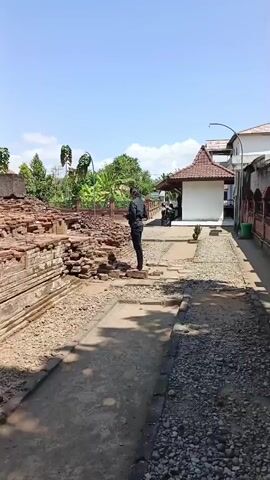 A man in a dark shirt and pants stands near an ancient stone structure under a clear blue sky. The structure is made of weathered bricks and stones, appearing to be ruins. In the background, there is lush greenery, trees, and some buildings with red tile roofs. The man is facing away from the camera and towards the ruins, with his hands in his pockets. The scene is outdoors, suggesting a historical site or archaeological dig.