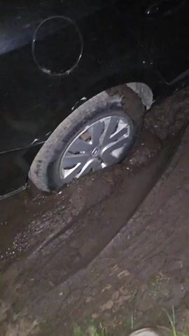 A black car is stuck in mud at night. A person is standing next to a trailer with a license plate. The person is looking at the trailer, and another person is holding up a phone. The car's tires are covered in mud.