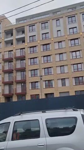 A slow panning shot from inside a vehicle shows a modern apartment building with beige brick cladding and dark-framed windows. The building has multiple stories with balconies featuring dark railings. In the foreground, the rear of a silver SUV is visible. The scene appears to be an urban environment under an overcast sky, with some construction barriers and trees also in view.