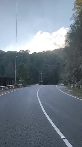 The video captures a view of a paved road winding through a mountainous, forested area. The road has a white line marking the center. On either side of the road are steep, rocky slopes covered in dense vegetation. There are some small structures or shelters visible near the side of the road in the distance. The sky is partially cloudy with sunlight breaking through. The camera appears to be moving along the road, suggesting a travelogue or scenic drive perspective. The overall environment is natural and rugged.