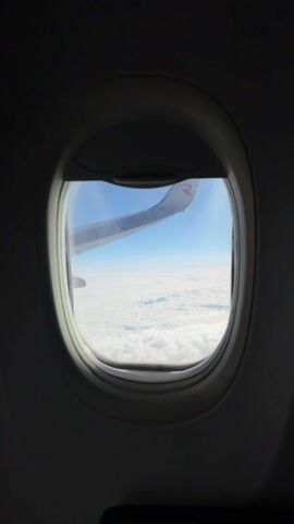 The video shows a view from an airplane window. Initially, the wing of the plane is visible with a red "R" logo. The camera then zooms out, focusing on the thick layer of clouds stretching across the horizon below a blue sky. The clouds appear dense and blanket-like, obscuring any view of the ground below. The lighting is bright, indicating a sunny day above the clouds.