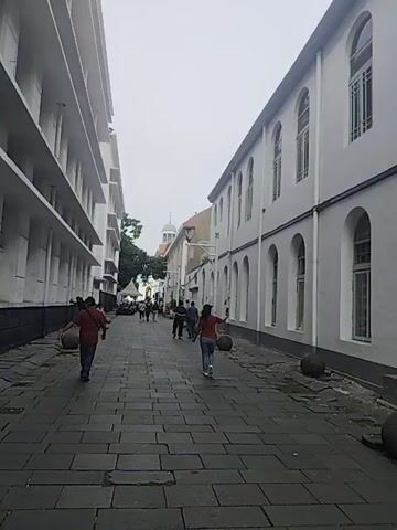 The video presents a street scene with a cobblestone-paved pedestrian walkway lined by multi-story buildings on either side. The buildings have white facades with repetitive window designs. Several people are walking in the same direction down the center of the walkway, some carrying items. The sky is overcast, suggesting a cloudy day. At intervals along the walkway are large, spherical stones resting on the edges.