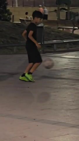 A child in a black t-shirt and shorts, wearing bright green soccer cleats, is playing soccer on a paved outdoor area at dusk. The child dribbles the ball with their right foot. In the background, other people are visible sitting on benches and standing near playground equipment. The lighting suggests it is evening.