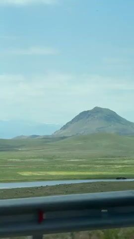 The video shows a landscape view from a moving vehicle. In the foreground, a silver metal guardrail is visible. Beyond the guardrail is a wide, green, and slightly undulating plain dotted with patches of pinkish-purple flowers. A body of water, possibly a lake or river, runs horizontally across the middle ground. In the background, a large, rounded mountain dominates the horizon under a pale blue sky with scattered clouds. The lighting suggests it is daytime, possibly late morning or early afternoon. The overall scene conveys a sense of open, rural, or undeveloped terrain.