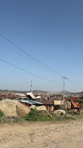 The video shows a dusty, unpaved road with several large, old dump trucks parked along the side. The trucks are various colors, including orange, yellow, blue, and white, and appear worn and heavily used. Some of the trucks are loaded with materials, potentially sand or gravel. In the background, there is a view of a town or village with buildings of varying sizes and styles, including what appears to be a church. Power lines run overhead, and a communications tower can be seen in the distance. The sky is clear and blue, suggesting a sunny day. A motorcycle with a passenger wearing a white veil drives past the trucks. Two people are visible sitting on the back of one of the orange and red trucks. The overall impression is of a busy working area in a developing location.