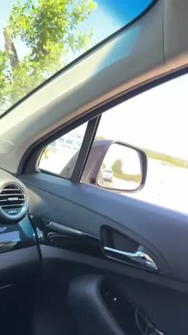 The video shows a view from inside a car driving on a sunny day. The dashboard of the car is visible in the foreground, with a digital display showing the time and other information. Outside the car, a road with a guardrail is visible, with construction work happening on the side. A lone tree stands in the middle ground, and in the distance, there are buildings and rolling hills under a clear blue sky with scattered clouds. The camera is angled upwards and to the side, capturing the sky and the landscape.