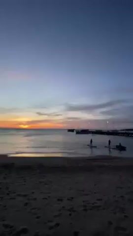 The video shows a sunset at a beach. The sky is filled with hues of blue, pink, and orange. The sun is setting over the horizon, casting a warm glow on the water. There are a few boats docked in the distance. Closer to the camera, several individuals are paddleboarding on the water. The beach is sandy and there are small waves lapping the shore.