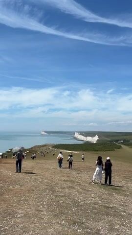 The video captures a panoramic view of a coastline with towering white cliffs. The sky is a vibrant blue, dotted with wispy white clouds. A vast expanse of calm, turquoise water stretches out towards the horizon. The foreground features a grassy, undulating hill with a clear path winding down towards the coast. Several people are scattered across the hill, some walking, others standing and observing the scenery. They are dressed in casual summer attire. The white cliffs, a prominent feature, are visible in the distance, forming a dramatic natural barrier against the sea. The overall impression is one of a popular tourist destination on a bright, sunny day, with people enjoying the scenic beauty of the coastal landscape.