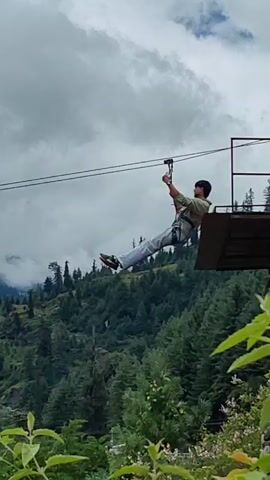 A person on a zip line is seen in the video. The person is wearing a harness and is holding onto the zip line. The background shows a lush green mountainous landscape with trees and clouds. The person appears to be enjoying the ride.