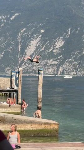 A person is performing a dive into a body of water from a wooden structure. Several other individuals are in the water, some partially submerged. In the background, a large mountain range is visible, with a boat on the water. The weather appears to be sunny.