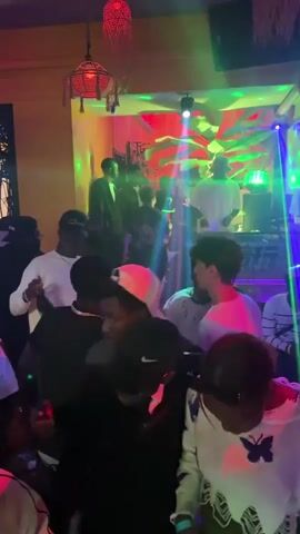 A medium-shot video taken in a nightclub shows the DJ and several clubgoers dancing. The DJ wears a white t-shirt and a dark baseball cap. He stands behind a console, manipulating the controls. Multicolored lights illuminate the room, flashing on and off to the beat of the music. The walls are decorated with vibrant, tropical-themed murals and hanging lanterns. The clubgoers are dressed casually and appear to be enjoying themselves. They dance, chat, and move around the room. The atmosphere is energetic and lively.