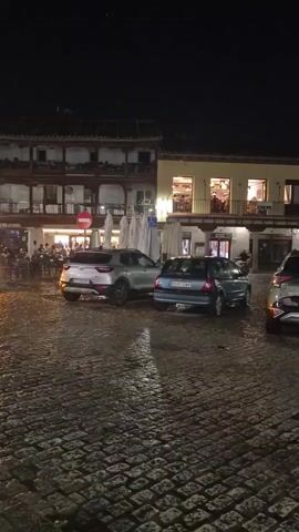 The video pans across a cobblestone plaza at night. Several parked cars are visible, including a silver SUV, a blue hatchback, a green SUV, a silver sedan, and a red hatchback. Behind the cars, buildings with illuminated windows line the plaza. The buildings appear to be restaurants or shops, with tables and chairs set up outside some of them. There are a few people visible in the background, walking and sitting at tables. A handicapped parking sign is visible near one of the buildings. The sky is dark, and it appears to be raining or recently rained, as the cobblestones are wet and reflective. A church steeple is visible in the distance.