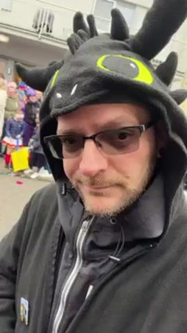 A man wearing a black dragon-themed hood with bright yellow eyes and small horns stands outdoors. He has glasses on, a beard, and is wearing a black fleece jacket and a layered black top underneath. The background is a street with a parade or festival going on, featuring people in costumes. There's confetti on the ground and buildings are visible in the background.