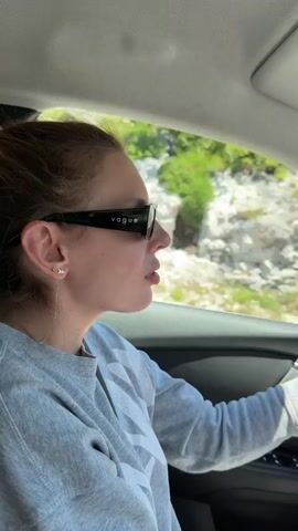 This video shows a woman driving a car on a sunny day. She is wearing sunglasses and a grey sweatshirt. The car is on a road with trees and rocks on either side. The video is filmed from inside the car, showing the driver's perspective.