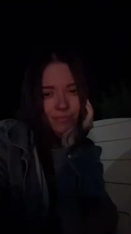The video begins with a close-up of a woman with long dark hair, wearing a denim jacket over a hooded sweatshirt. Her hand is resting on her cheek as she looks around and then directly at the camera, smiling slightly. The background is dark and blurry. The camera then pulls back to reveal a group of people gathered around a bonfire at night. One person is standing and gesturing. The fire is burning brightly, illuminating the faces of the people sitting and standing around it. The environment appears to be outdoors, possibly a backyard or campground, with a structure and trees visible in the background. The overall mood seems relaxed and social.