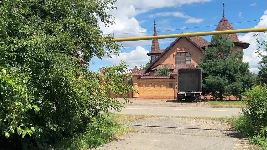 This media file depicts an outdoor scene on a sunny day with some clouds. The camera is positioned low, looking across a paved area towards a large, multi-story house with distinctive conical turrets and a brick fence. The house appears to be made of brick with a brown roof. In front of the house, a large delivery truck with its rear doors open is parked on the road.

The foreground is dominated by lush green bushes and trees, with a yellow pipe running horizontally across the frame, supported by metal poles. The pipe appears to be a gas line. The scene suggests a residential area in a rural or suburban setting, possibly in Dondukovskaya, Russia, given the context. The vegetation is thick and green, indicating a warm season, likely summer. The sky is partly cloudy, with some large white cumulus clouds against a blue sky. The overall mood is tranquil and ordinary, with the presence of the truck suggesting a delivery or move is in progress. No people are visible in the frame. There is no visible text in the media file.