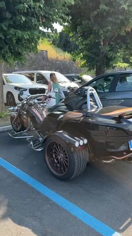 A woman with blonde hair, wearing a green dress and carrying a white tote bag, stands near a trike motorcycle. The trike is dark brown with copper accents, large rear wheels, and chrome accents. It is parked in a lot with a blue line on the asphalt. Several cars, including a white SUV and a gray sedan, are parked nearby. Trees with green leaves provide shade. A stone wall is visible in the background.