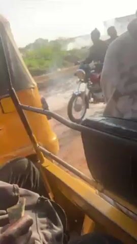 The video shows a view from inside a yellow auto-rickshaw moving on a muddy, dirt road. Another motorcycle with two people on it drives past in the opposite direction. Smoke is visible in the background. The person holding the camera is holding some currency.