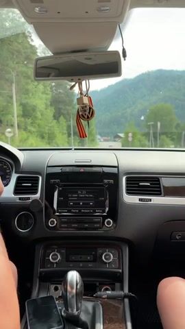 This media file shows the interior of a car, specifically the dashboard and the view through the windshield. The camera appears to be mounted on the dashboard, facing forward.

**Subjects:**
*   **People:** The legs of two individuals are visible, one on the left and one on the right, suggesting they are passengers or the driver and a passenger in the front seats. The hand of the driver is also visible on the steering wheel.
*   **Objects:** Prominent objects include the car's dashboard, steering wheel, gear shifter, car stereo system, rearview mirror, a hanging air freshener with a St. George's ribbon, and a smartphone plugged into a charging port.
*   **Scene/Setting:** The scene is the interior of a car driving on a paved road. Outside the car, a rural or mountainous landscape is visible, characterized by green trees, hills or mountains, and some distant buildings. The sky appears overcast.

**Activity/Event:**
The primary activity is driving. The car is in motion, and the view through the windshield shows a road winding through a natural landscape. The car stereo is playing music, as indicated by the display.

**Notable Details:**
*   **Time of Day:** The lighting suggests it is daytime, possibly late morning or afternoon, given the overcast conditions.
*   **Weather:** The sky is cloudy, indicating overcast weather.
*   **Location Cues:** The presence of a St. George's ribbon, a symbol commonly associated with Victory Day celebrations in Russia and other post-Soviet states, and the general landscape are consistent with a location in Russia, such as Uznezya. Road signs are also visible in the distance.
*   **Emotions:** The imagery conveys a sense of travel and perhaps a relaxed atmosphere, typical of a road trip.

**Visible Text:**
The car stereo display shows the following text:
*   "10:23" (Time on the stereo)
*   "CC Catch - Cause You Are..." (Song title and artist)
*   "1:10" and "-3:10" (Track progress indicators)
*   Buttons with labels such as "RADIO", "MEDIA", "PHONE", "CAR", "SETUP", and "LIST" are visible on the stereo.
*   A speedometer is visible on the dashboard, showing a speed of approximately 70 km/h.