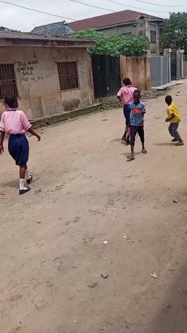 The video shows a street scene with several children playing. In the foreground, a young boy wearing a blue t-shirt and blue jeans stands with his back to the camera. To his left, closer to the camera, another child is partially visible. In the middle ground, a group of children are playing. Three children in pink shirts and blue skirts are walking away from the camera towards a building on the left. Two boys are engaged in activity in the center of the street, with one boy in a blue and white shirt and another in a yellow shirt. Further down the street, another child in a pink shirt and blue skirt is walking away from the camera. The environment appears to be a dirt road with buildings on either side. One building on the left has graffiti on its wall. The sky is overcast. The overall impression is a casual street game among children.