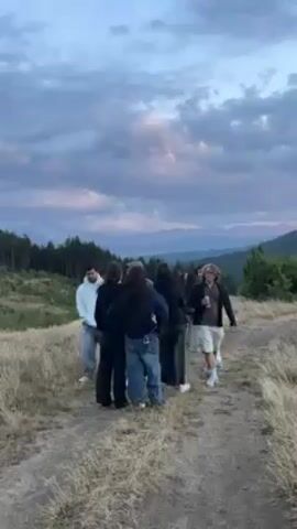A group of approximately ten young people stand on a grassy hill in a wooded area. They are arranged in a semi-circle, looking out at a distant mountain range under a cloudy sky at dusk. One individual in a light-colored sweatshirt and jeans stands slightly apart from the group. The ground is covered in dry grass, and a dirt path runs near the group. The lighting suggests it is late afternoon or early evening.