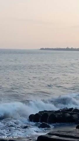 The video shows a rocky shoreline with waves crashing against the rocks. The sky is overcast and the water is a calm blue. In the distance, there is a line of trees and buildings. A single person is standing on the rocks, looking out at the sea. The camera is positioned at eye level, with a slight downward angle. The video is of good quality, with clear images and sound.