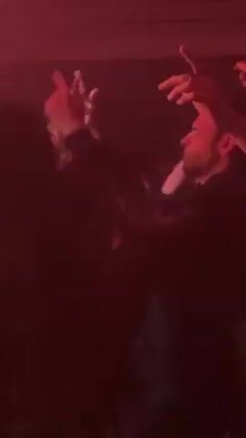 The video, bathed in red light, shows a crowded indoor space. People are moving and obscured by the dim lighting. One person in the foreground is wearing a black long-sleeved shirt with a white design on the front. The red light and blurred figures suggest a concert or club setting.