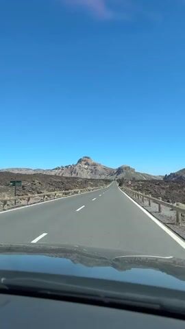 The video shows a first-person perspective from a car driving on a paved road through a volcanic landscape under a clear blue sky. The road has a white line down the center and a guardrail on the right side. The landscape on either side of the road is rocky and barren, with some low-lying vegetation. In the distance, mountains can be seen. There are no people or other vehicles visible in the video. The sun is shining, casting shadows.