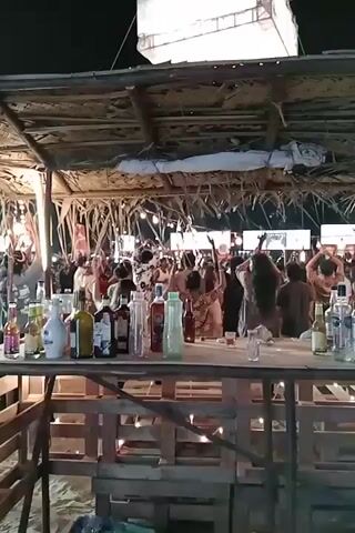 A video depicts a night scene at a beachside bar with a large group of people dancing in the background. The bar itself is made of wooden pallets and has a thatched roof. Numerous bottles of alcoholic beverages, including clear and dark liquids, are displayed on the bar top. Small, warm-toned string lights are draped around the wooden structure of the bar. In the background, under another thatched structure, a crowd of people is actively dancing, their arms raised in the air. A large white rectangular structure, possibly a tent or screen, is visible in the upper background against the dark night sky. The ground appears to be sand.
