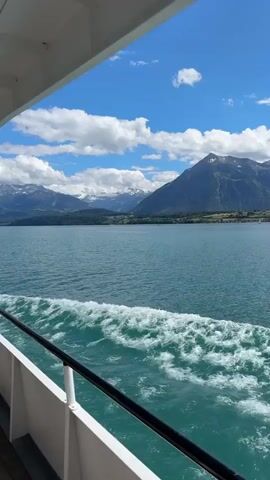 The video shows a view from the side of a white boat moving through a large body of turquoise water. The boat has a white railing with a black handrail along the side. White foam and waves are visible trailing behind the boat in the water. In the distance, there is a shoreline with green hills and trees, and behind that, large, majestic mountains, some of which are capped with snow. The sky is a bright blue with scattered white clouds. The overall scene is serene and natural, with a clear emphasis on the vastness of the water and the grandeur of the mountains.
