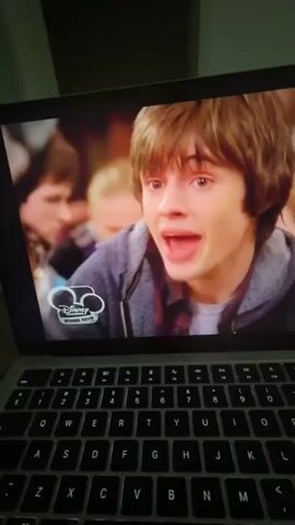 The video shows a laptop screen displaying a paused scene from a television show. On the screen, a young man with brown hair is looking intensely at the camera, his mouth open in an expression of surprise or anger. He is wearing a gray hooded sweatshirt over a plaid shirt. A Disney Channel logo is visible in the lower left corner of the screen. The laptop's keyboard is partially visible in the foreground, indicating the video is being watched on a computer. In subsequent frames, a young woman with blue eyes and a brown hat is shown holding a blue iPod, and another young man with glasses and a fedora hat appears. The scene appears to be indoors, with other people blurred in the background.