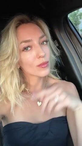 A blonde woman with her hair down is sitting in a car. She is wearing a black strapless top and a gold necklace with a heart pendant. She touches her hair and looks at the camera with her eyes closed. Then she opens her eyes and looks to the side.