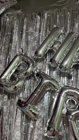 The video shows a close-up shot of silver foil balloons arranged to spell out "HAPPY BIRTHDAY". The balloons are hanging against a backdrop of silver tinsel curtains. Some of the balloons are slightly deflated, and the lighting is dim. There are also a few star-shaped balloons visible at the top of the frame. The overall impression is of a party decoration.