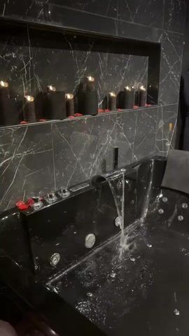 A camera pans across a dimly lit, luxurious room. The room appears to be a spa or a romantic getaway suite. The first object in view is a large, black jacuzzi tub, with water filling it. The tub has several built-in jets and two headrests. The walls behind the tub are covered in dark marble with a shelf containing several black candles and red rose petals. To the right of the tub, a black robe hangs on a hook. The camera continues to pan, revealing a king-sized bed covered in a gray comforter and decorated with red rose petals and black pillows. Two bedside tables with golden lamps flank the bed. A path of lit white candles leads from the bed across a dark rug on a wooden floor. Further to the right is a massage table covered in red upholstery and scattered with rose petals. A black chair and a tall black cabinet containing what appears to be shoes and other items are also visible. The room is furnished with dark, elegant decor, creating a mood of intimacy and relaxation.