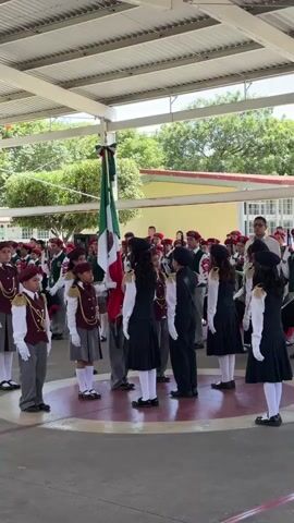 A group of children in uniform are gathered outdoors, around a flagpole bearing the Mexican flag. The children are dressed in formal attire with maroon berets, vests over white shirts, and some are wearing skirts while others wear pants. The flag is partially raised on the pole. The event appears to be a flag-raising ceremony, possibly at a school. The background shows a building with yellow walls and a white roof.