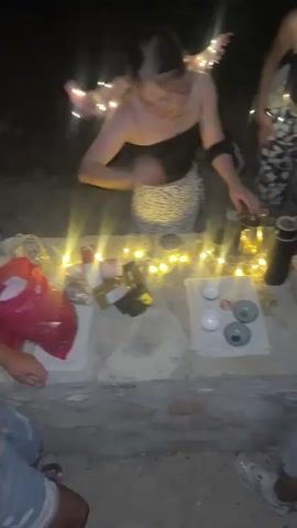 The video opens on a nighttime scene, illuminated by string lights. Four people are gathered around a stone surface. In the foreground, a young woman with blonde hair pulled back is leaning over the surface, holding a small white cup and appearing to pour liquid from a dark thermos. To her right, a person wearing glasses and a dark top with string lights is standing. To the left and right of the woman, partially visible, are other individuals. On the stone surface, there are several small cups, some containing liquid, and various bottles and containers. The background is dark, suggesting an outdoor setting. The scene transitions to other views of a nighttime gathering, with more people visible, some near what appears to be a grill, and others walking or standing. String lights are prominent throughout, creating a festive atmosphere.