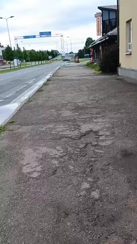 A first-person perspective video walks along a broken concrete sidewalk. A two-lane road with wet pavement runs parallel to the sidewalk. Buildings line the other side of the road, including a building with a red sign that reads "PENOK". Overhead signs and traffic lights are visible in the distance. The sky is overcast.
