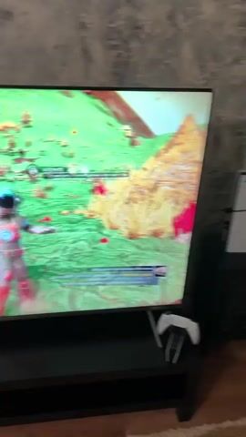 The video shows a person playing a video game. The video begins with a TV screen displaying a game featuring two characters in spacesuits walking on a green landscape. The camera then pans to reveal a person sitting at a desk, wearing a red and blue patterned robe, using a keyboard and mouse to play the game displayed on the monitor. The desk also contains a keyboard, a mouse, paper, and a pen. The room has wood flooring and blue patterned wallpaper.