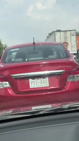A woman is seen standing next to a red car on a highway. She is wearing a pink shirt and blue jeans. She opens the driver's side door of the red car and gets into the driver's seat. The video is shot from inside another vehicle, looking forward. The sky is cloudy. Other vehicles are visible on the highway, including a white truck and an orange shipping container.