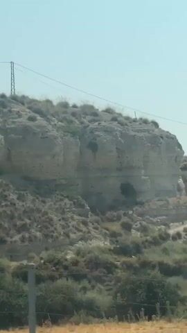 The video shows a moving shot of a rocky, arid hillside. The hillside is covered in sparse vegetation, mostly dry grasses and scrub bushes. In the upper part of the frame, a tall, thin electrical pylon with wires stretching across the sky is visible. The rock formations appear layered, indicating geological strata. The lighting suggests it is daytime with a clear sky. The camera is moving from left to right, giving the impression of traveling past the hillside.