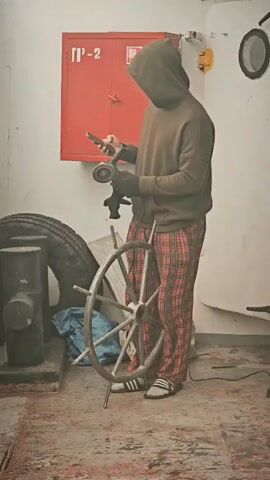 A person wearing a brown hooded sweatshirt and red plaid pajama pants stands holding a grinder in their right hand and a smartphone in their left hand. The person is facing away from the camera. To the left of the person is a large tire and a metal post. In the center of the frame is a ship's wheel. Behind the person is a red metal cabinet with Cyrillic writing. The floor is dirty and stained.