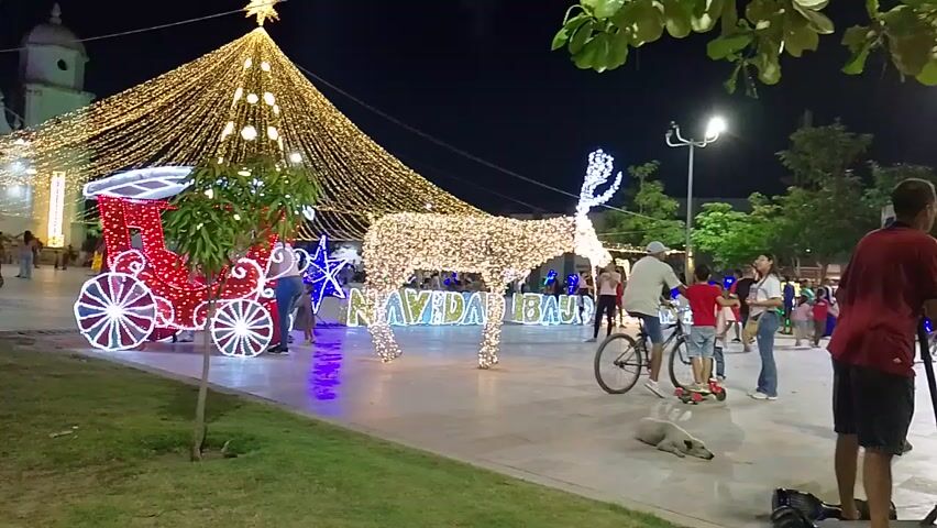 At night, a town square is decorated with Christmas lights. A large light display of a Christmas tree hanging from overhead wires is to the back, with a star on top. In front of the tree is a Christmas carriage light display. To the right is a reindeer light display spelling out "Navidad Baure." People are milling around the square. A person rides a bicycle by the reindeer display, and a boy stands on a scooter next to them. A dog lies on the ground near the scooter.